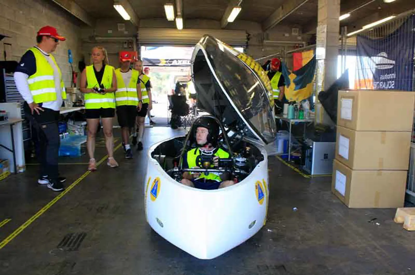 Solbilen Nova Lumia i garaget i Hidden Valley, Darwin, med medlemmarna i JU Solar Team 2025 stående runt den och diskuterande.